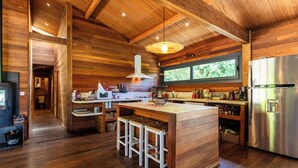 A modern kitchen with wooden walls, a central island with stools, stainless steel appliances, and a large window providing natural light. - House / Villa - Porto vecchio (Porto-Vecchio)