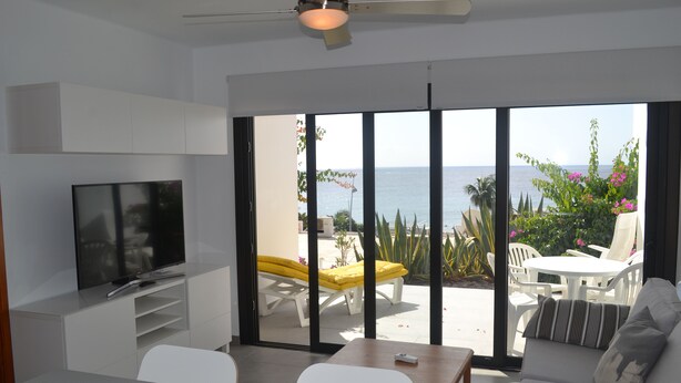 Een moderne woonkamer met een flatscreentelevisie, een bank, een eettafel en uitzicht op zee door grote glazen deuren.