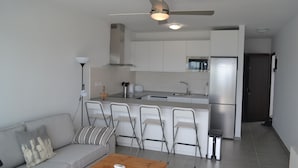 Een compacte keuken met een ingebouwde magnetron, een roestvrijstalen koelkast en een klein eiland met een spoelbak. De woonruimte heeft een bank met een salontafel en vier stoelen.