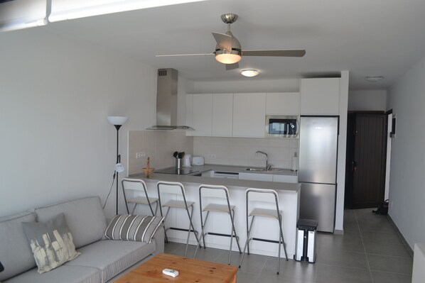Cocina compacta con microondas integrado, refrigerador de acero inoxidable y una pequeña isla con pileta. En la zona de estar, hay un sofá, una mesa de centro y cuatro sillas.