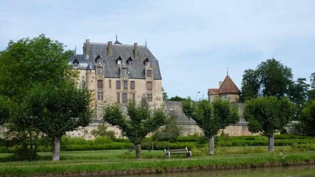 Un bâtiment historique doté d’un fossé, entouré d’arbres et bordé d’une étendue d’eau paisible.