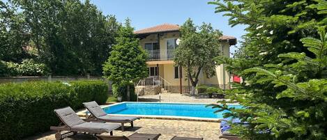 A pool area with lounge chairs, a clear blue pool, and a house with a balcony in the background.
