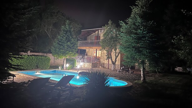 Vista nocturna de una casa con piscina, rodeada de árboles y un muro de piedra.