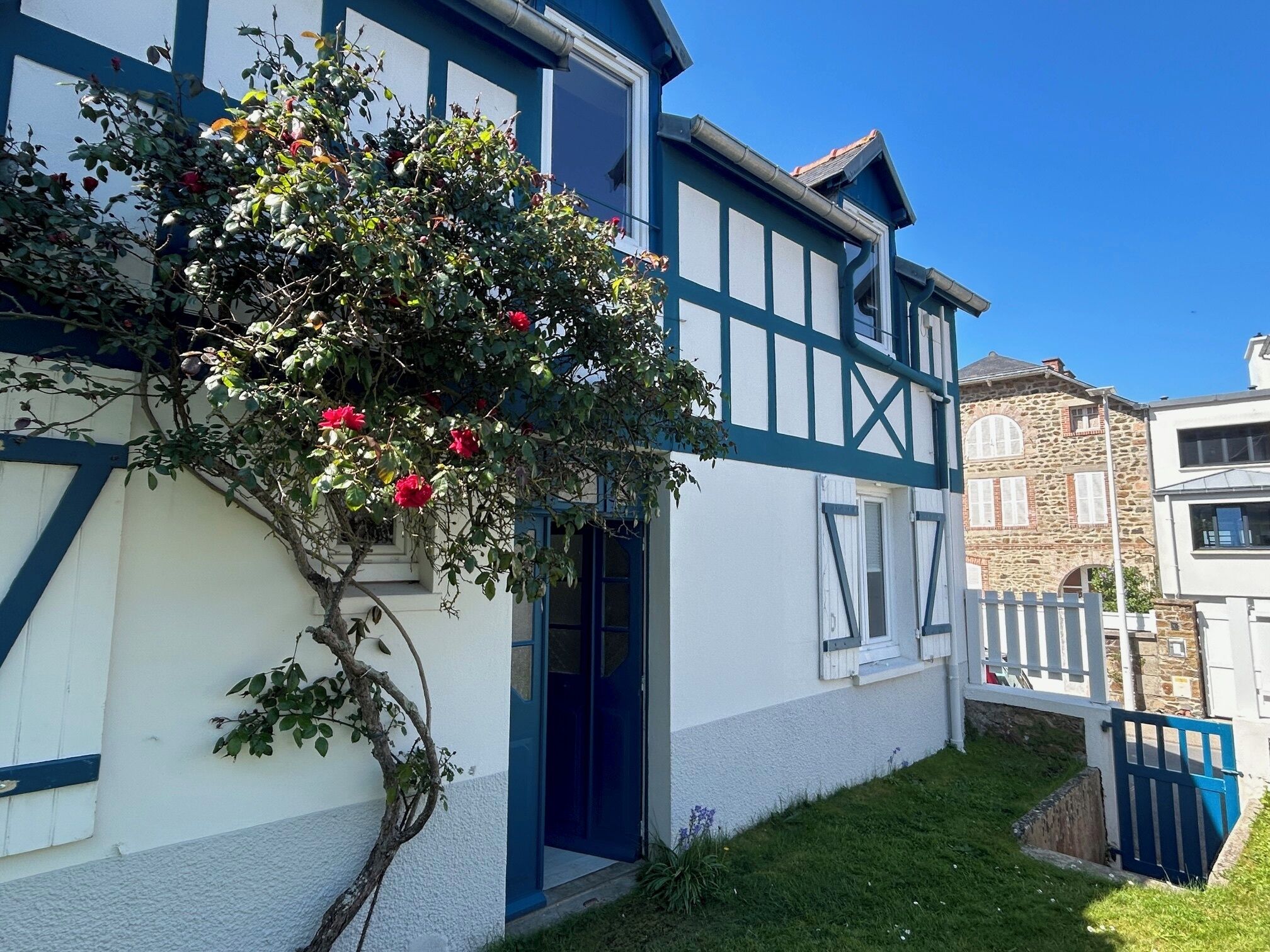 A traditional half-timbered house with a white facade and blue accents, a small garden with a flowering tree, and a stone building in the background.