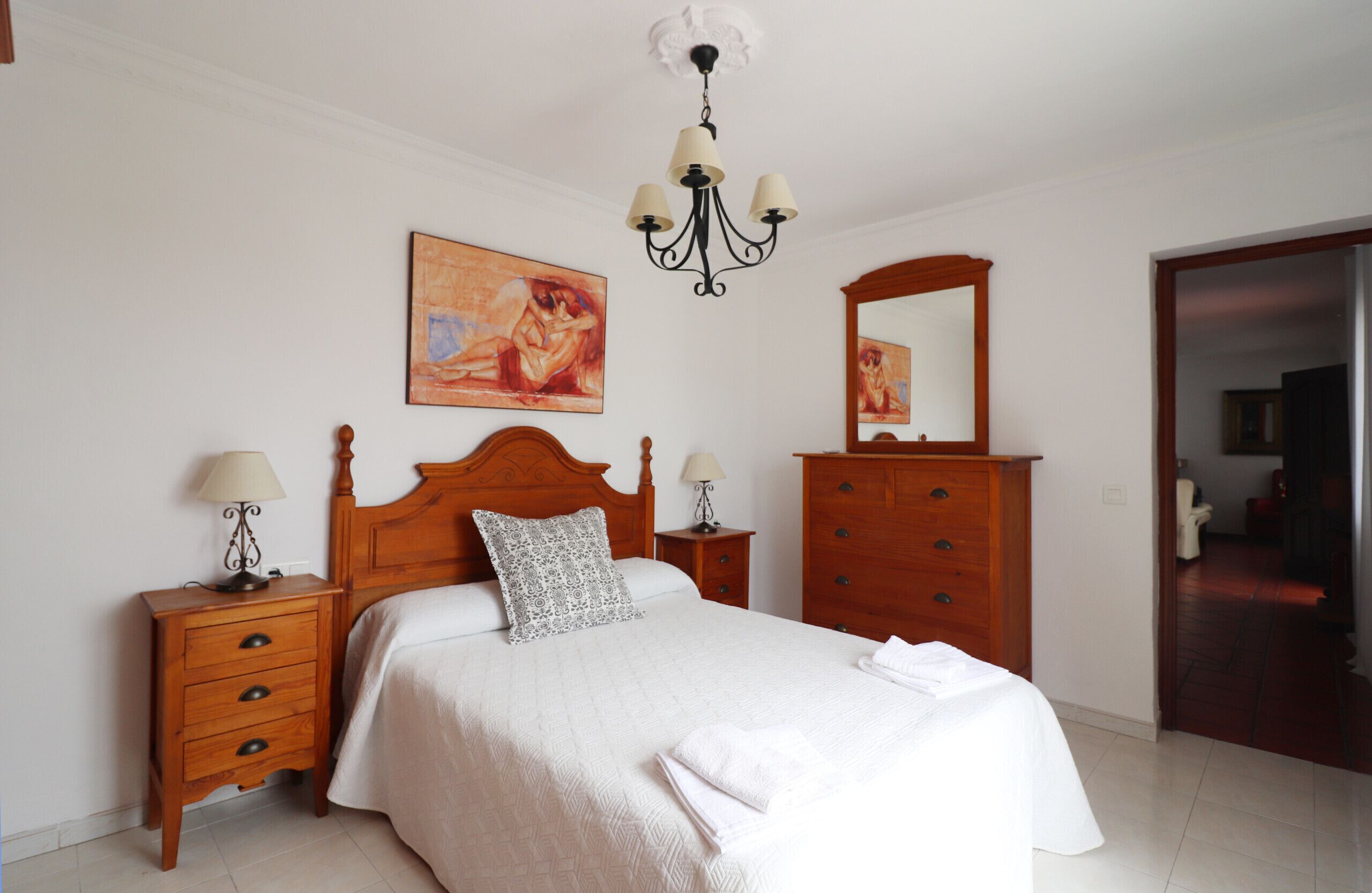 Een slaapkamer met een houten bed, twee nachtkastjes, een kroonluchter, een spiegel en een schilderij aan de muur.