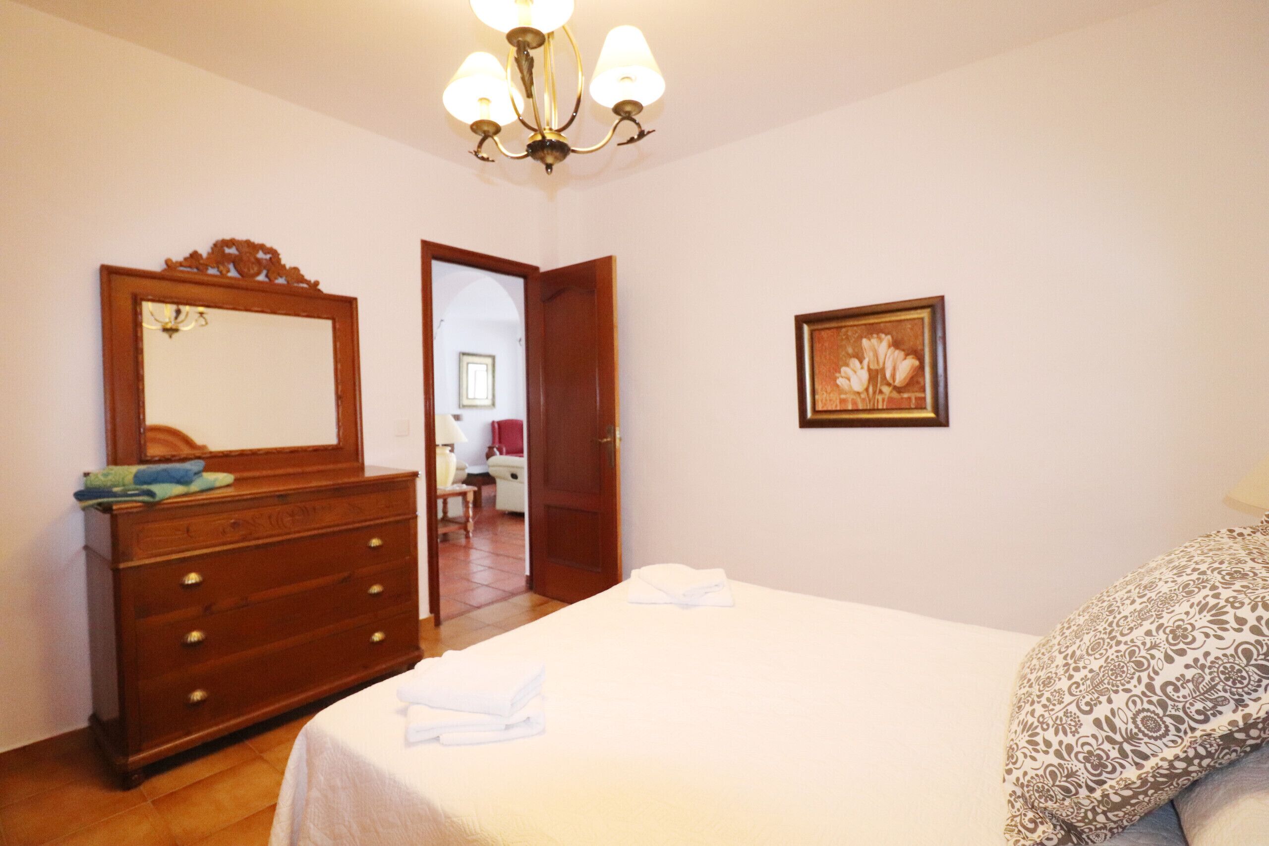 A bedroom with a wooden dresser, a bed with a patterned pillow, a framed picture on the wall, and a chandelier.