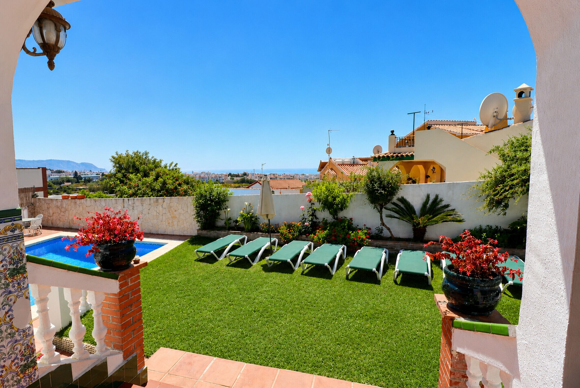 A poolside area with green lounge chairs, a small pool, and a view of the sea and buildings in the distance.