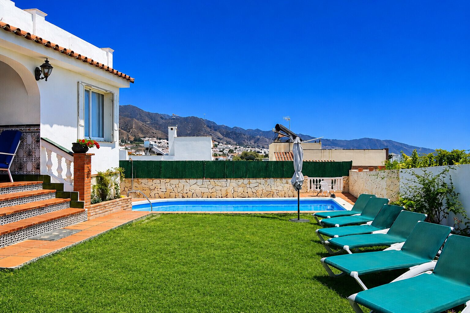 A pool area with a clear view of mountains, a set of stairs leading to a door, and a row of green lounge chairs.