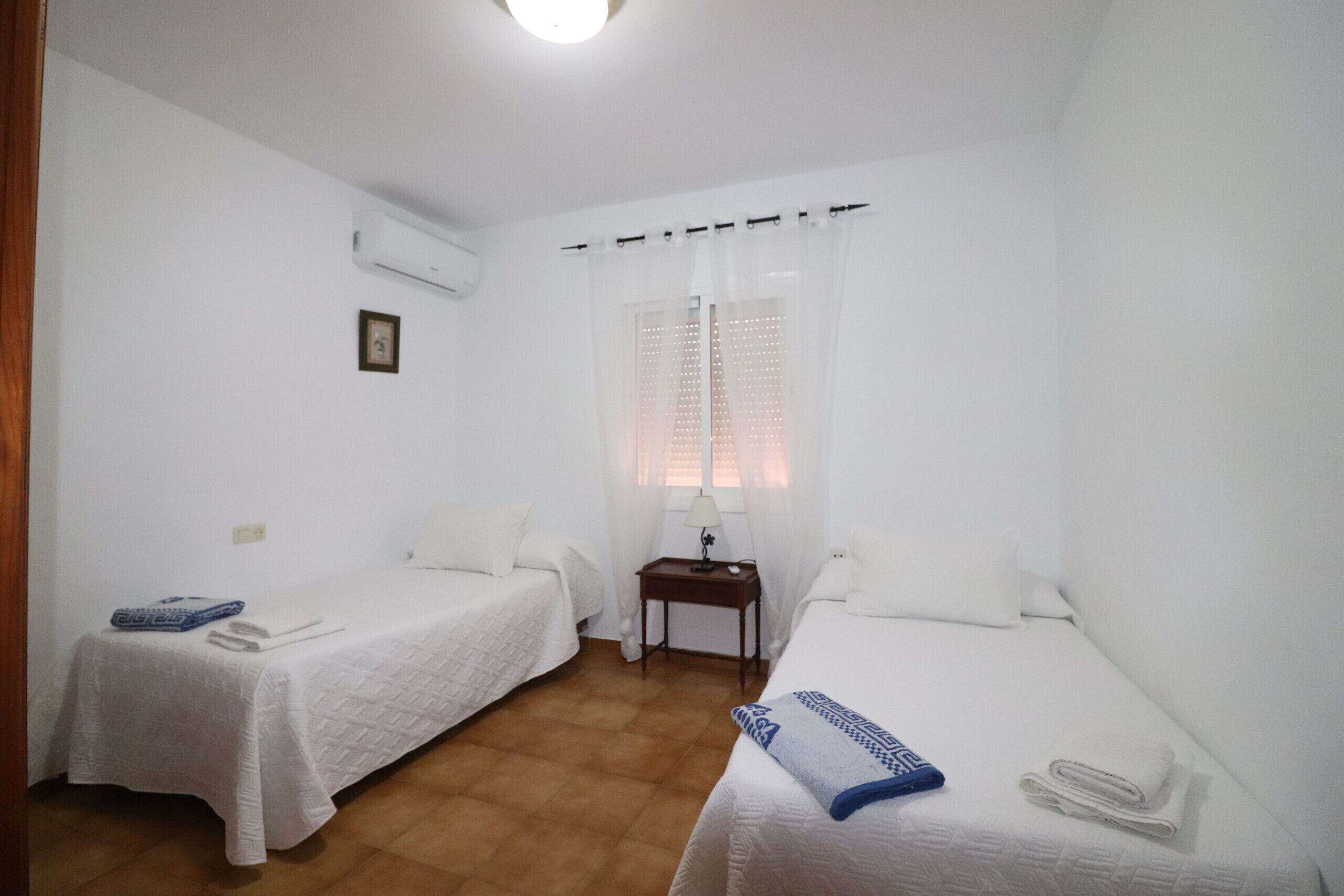 Una stanza piccola e pulita con due letti singoli, un comodino con lampada e una finestra con tende trasparenti.