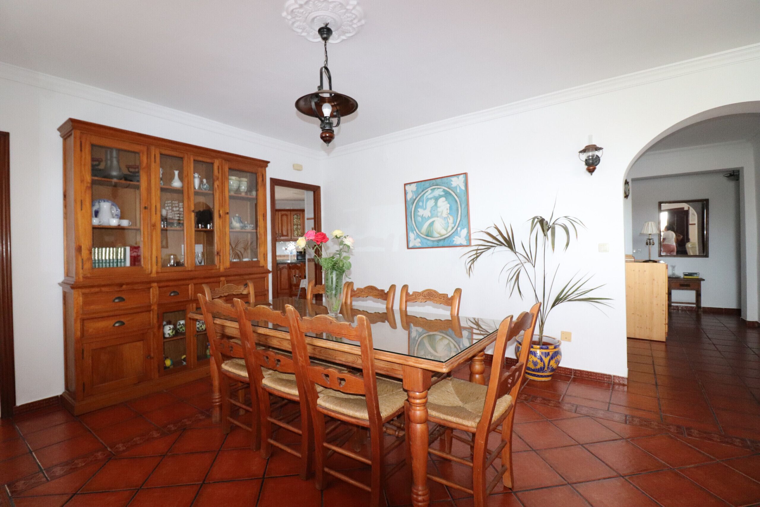 Un'area pranzo con un tavolo e sedie in legno, un mobile con vetrine, un vaso con fiori e un quadro appeso al muro.