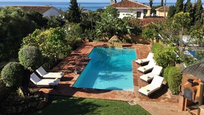 Une piscine rectangulaire bordée de pierres, entourée de chaises longues et d’un jardin bien entretenu où poussent différentes plantes et arbres.