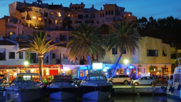 Un port de plaisance avec des bateaux amarrés, des palmiers et des bâtiments dont les enseignes sont illuminées en arrière-plan.