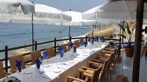Un espace repas avec des tables préparées pour deux personnes, offrant une vue sur la mer, agrémenté de parasols blancs et de chaises en osier.