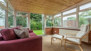 Une véranda avec un plafond en bois, de grandes fenêtres, un canapé rouge, un fauteuil à bascule blanc et une vue sur un jardin.