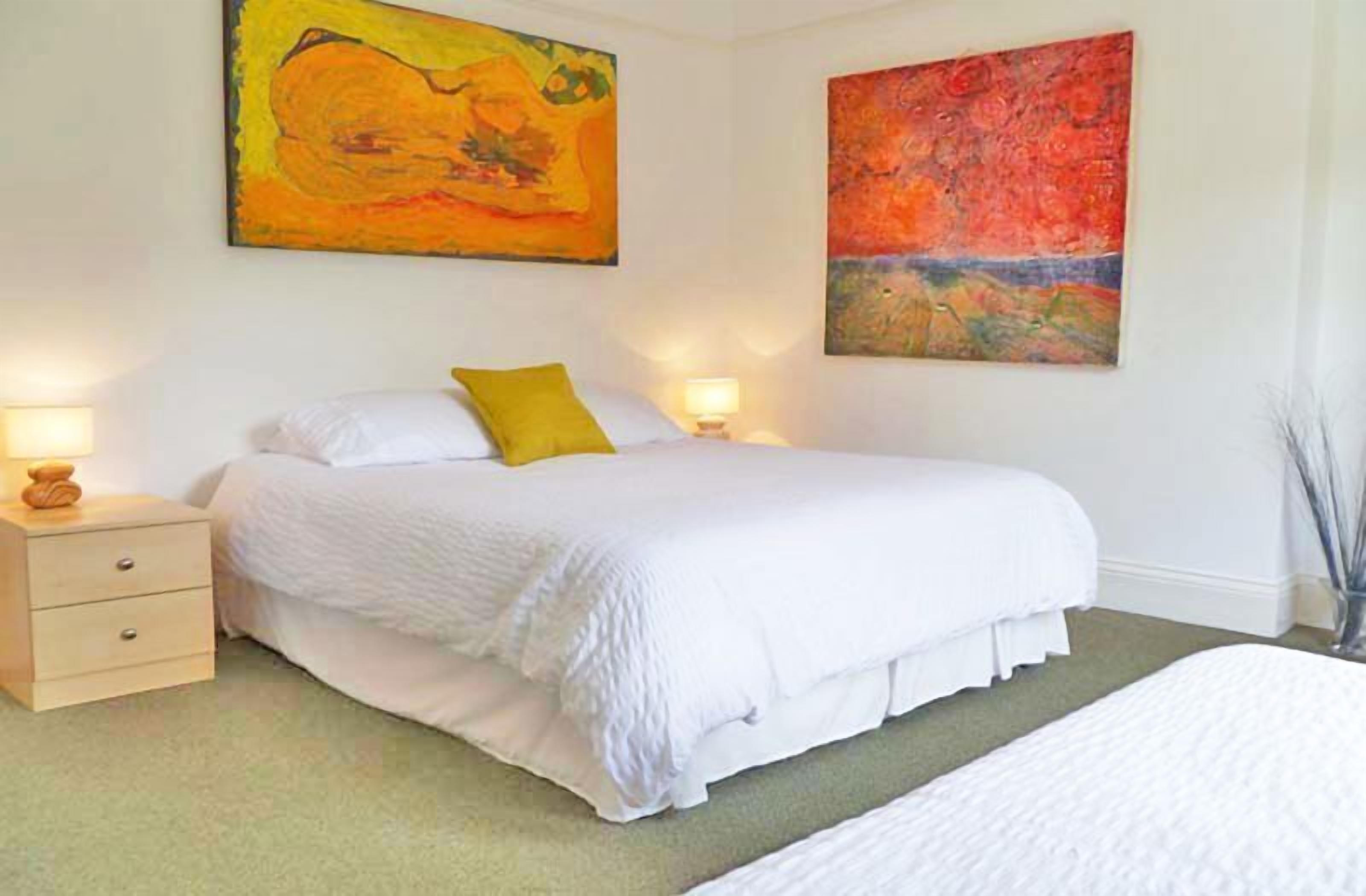 A bedroom with a large bed, two bedside tables with lamps, and two abstract paintings on the walls.