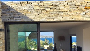 Un patio moderno al aire libre con paredes de piedra, un ventanal de vidrio que da a un comedor y vistas al mar.