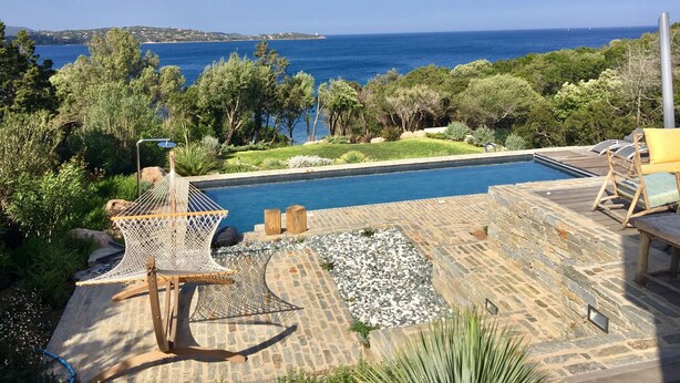 Zona de piscina con vistas al mar, rodeada de vegetación y suelo de piedra.