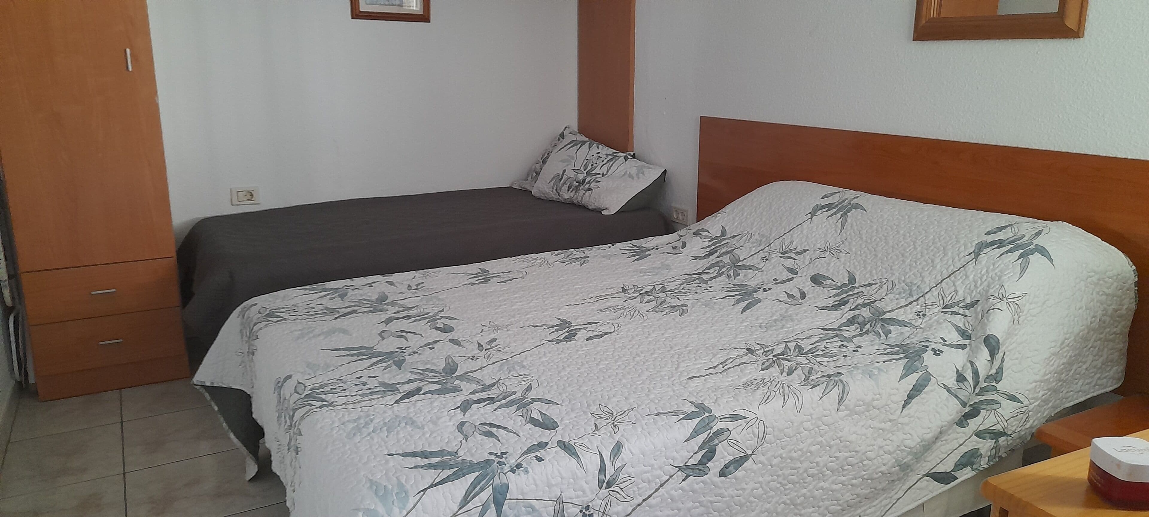 A bedroom with two beds, a wooden wardrobe, and a small table with a red object on it.