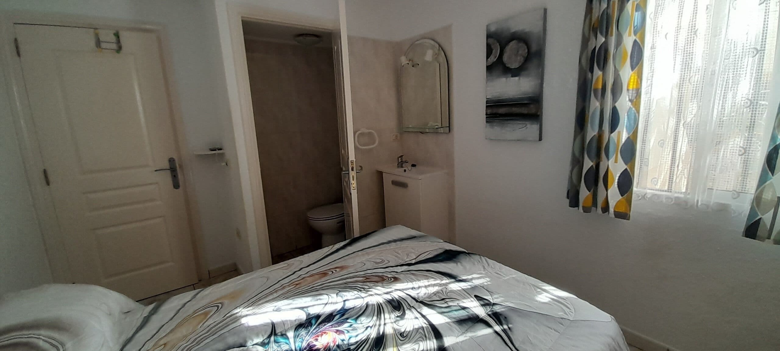 A bedroom with a bed, a bathroom with a toilet and sink, and a door leading to another room.