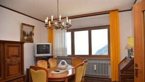 A dining area with a wooden table, chairs, a chandelier, a television, and a window with curtains. - Sagui apartment in the heart of the Dolomites CIN 025069C2H8ECUEQQ  (Zoppe' di Cadore (Belluno))
