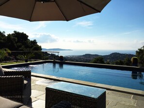 Una piscina infinita con vistas al mar y a las montañas, rodeada de muebles de mimbre y plantas en macetas.