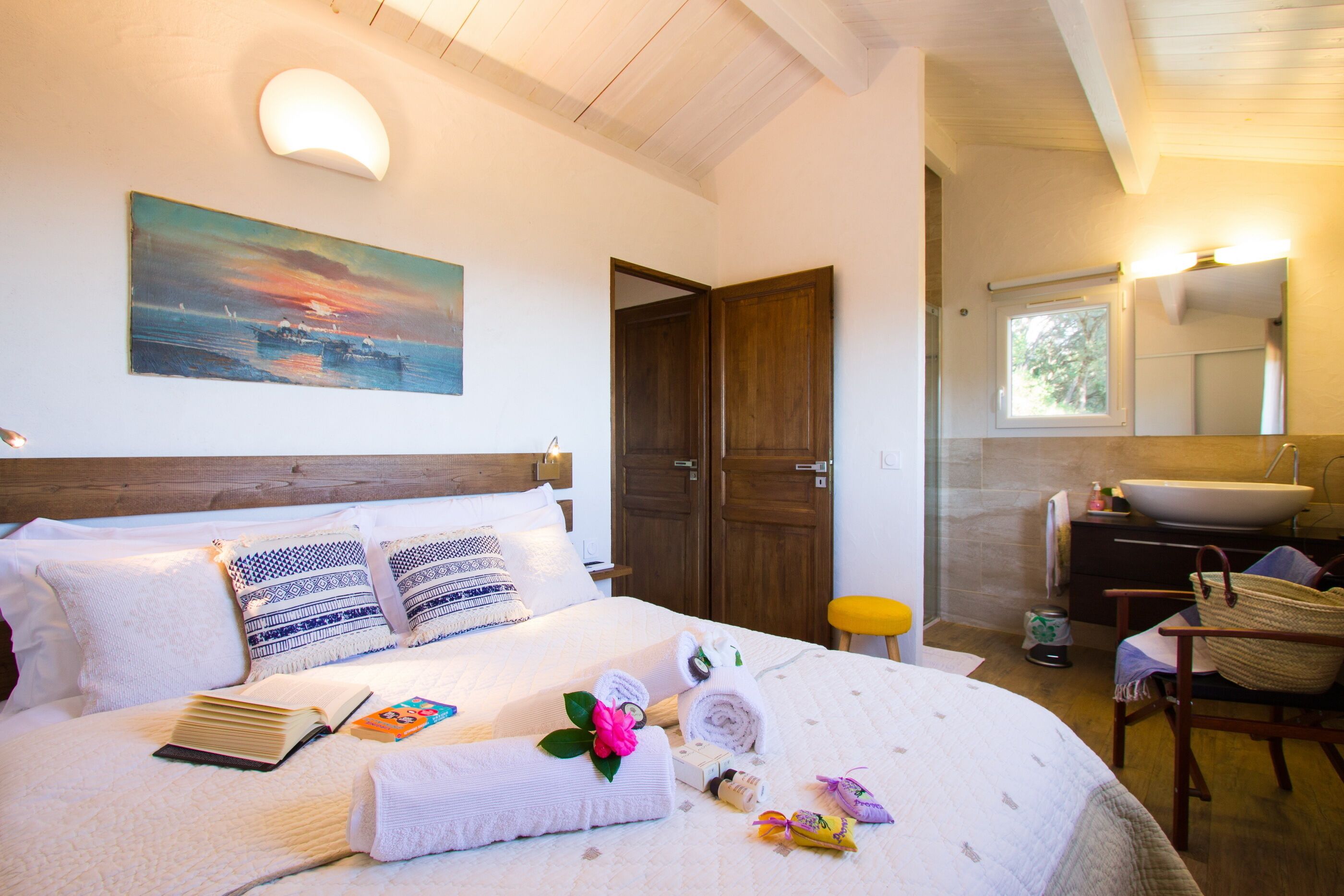 Une chambre à coucher avec un lit, des oreillers, une table de chevet avec des livres, un tableau, une salle de bain avec un lavabo et une chaise.