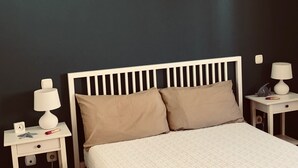 Un dormitorio con una cama blanca, dos almohadas color beige, una mesita de noche blanca con una lámpara y un cajón pequeño.