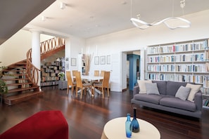 A spacious living area with a staircase, a dining table with chairs, a sofa, and a bookshelf filled with books.
