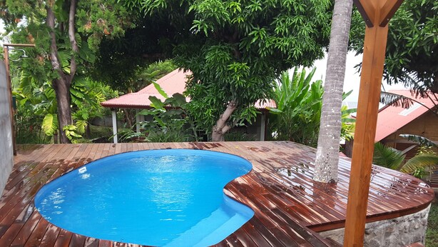 A swimming pool with a wooden deck, surrounded by lush greenery and a red-roofed structure. - Charming home comfort (LE PITON SAINT LEU)