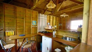 A compact kitchen with wooden walls, a small refrigerator, a sink, and a dining area with metal chairs. - Charming home comfort (LE PITON SAINT LEU)