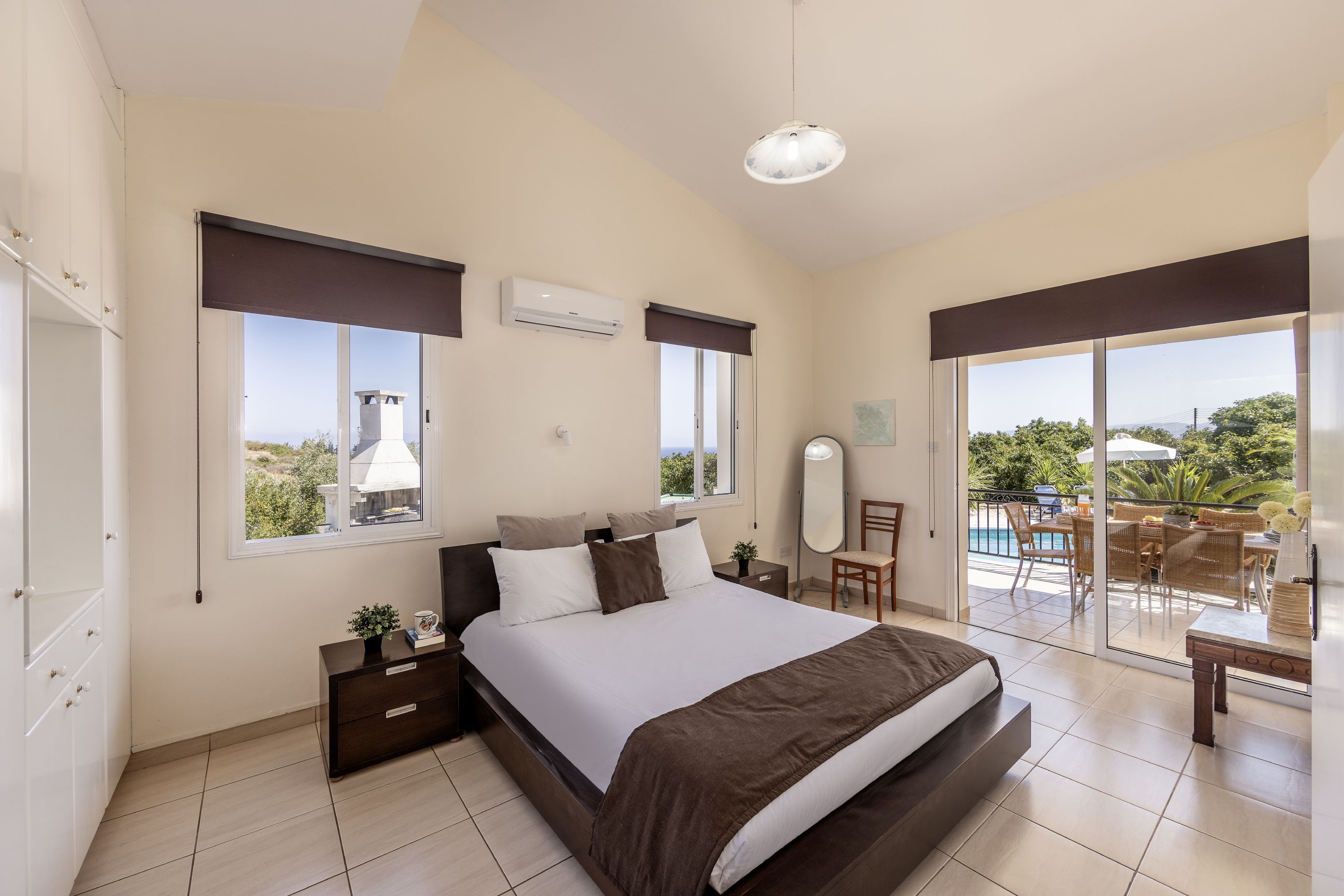 A bedroom with a bed, bedside tables, a nightstand, a window, an air conditioning unit, a mirror, and a view of the outdoors.