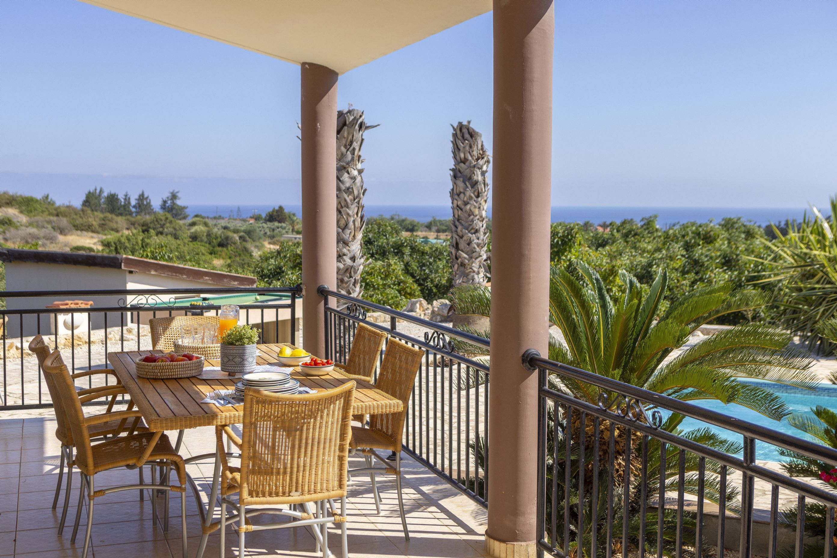 A terrace with a table set for dining, surrounded by wicker chairs, overlooking a pool and a view of the sea.