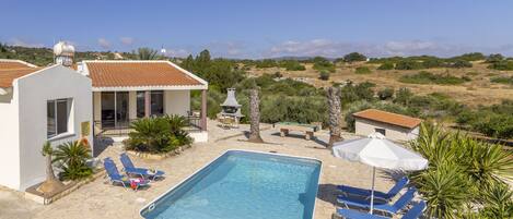 A swimming pool with lounge chairs and a table, surrounded by a garden and a view of the countryside.