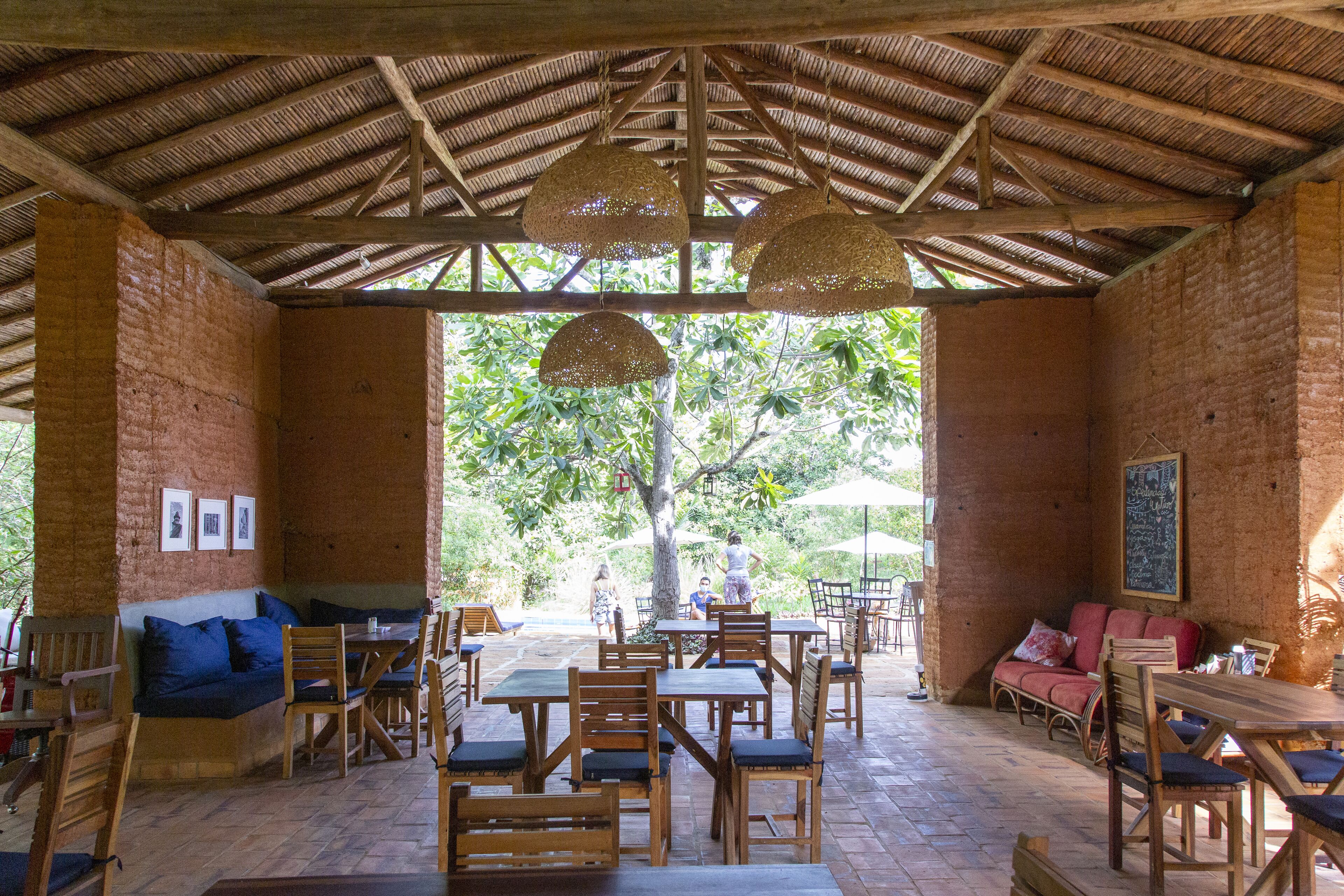An open-air dining area with wooden tables and chairs, a cushioned bench, and hanging lamps.