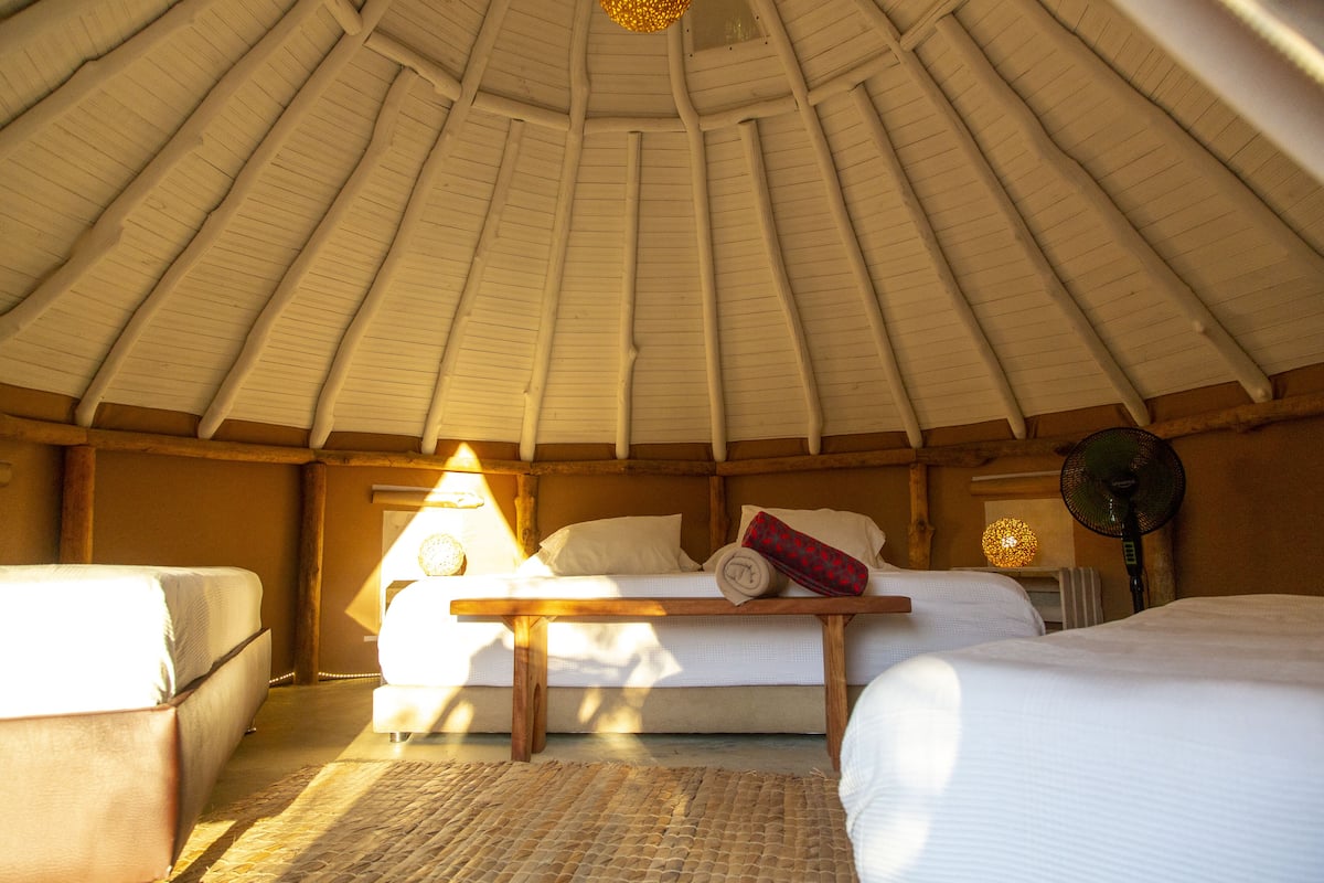 Een knus ogende kamer met een hoge, schuine kap, een bed met kussens, een nachtkastje en een lamp.