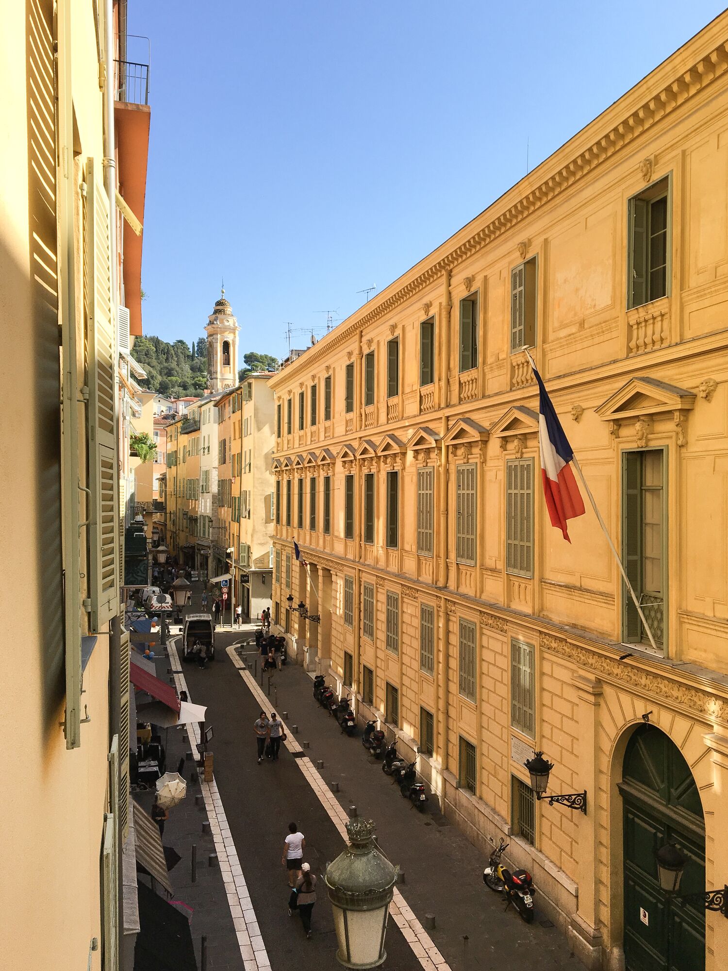 Vista d'un carrer amb un edifici que destaca per una gran bandera, una torre campanar i un passeig pavimentat.