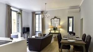 A modern living room with a sofa, dining table, and chairs. There is a TV on a stand, a chandelier, and a mirror above the dining table. - Spacious 3BR / 3BA Apartment in Old Town Nice • 2-Min Walk to Beach • Full A/C (Nice)