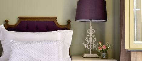A neatly made bed with a wooden headboard, a white pillow, and a decorative lamp with a purple shade on a bedside table.