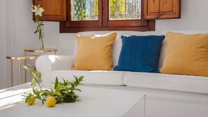 Un sofá blanco con dos cojines, una mesita con un florero y una ventana con contraventanas de madera.