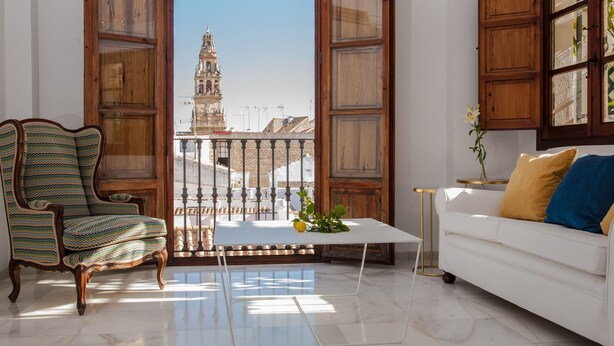 Un salón con vistas a un edificio histórico, un sofá blanco, un sillón estampado y una mesita con una planta.
