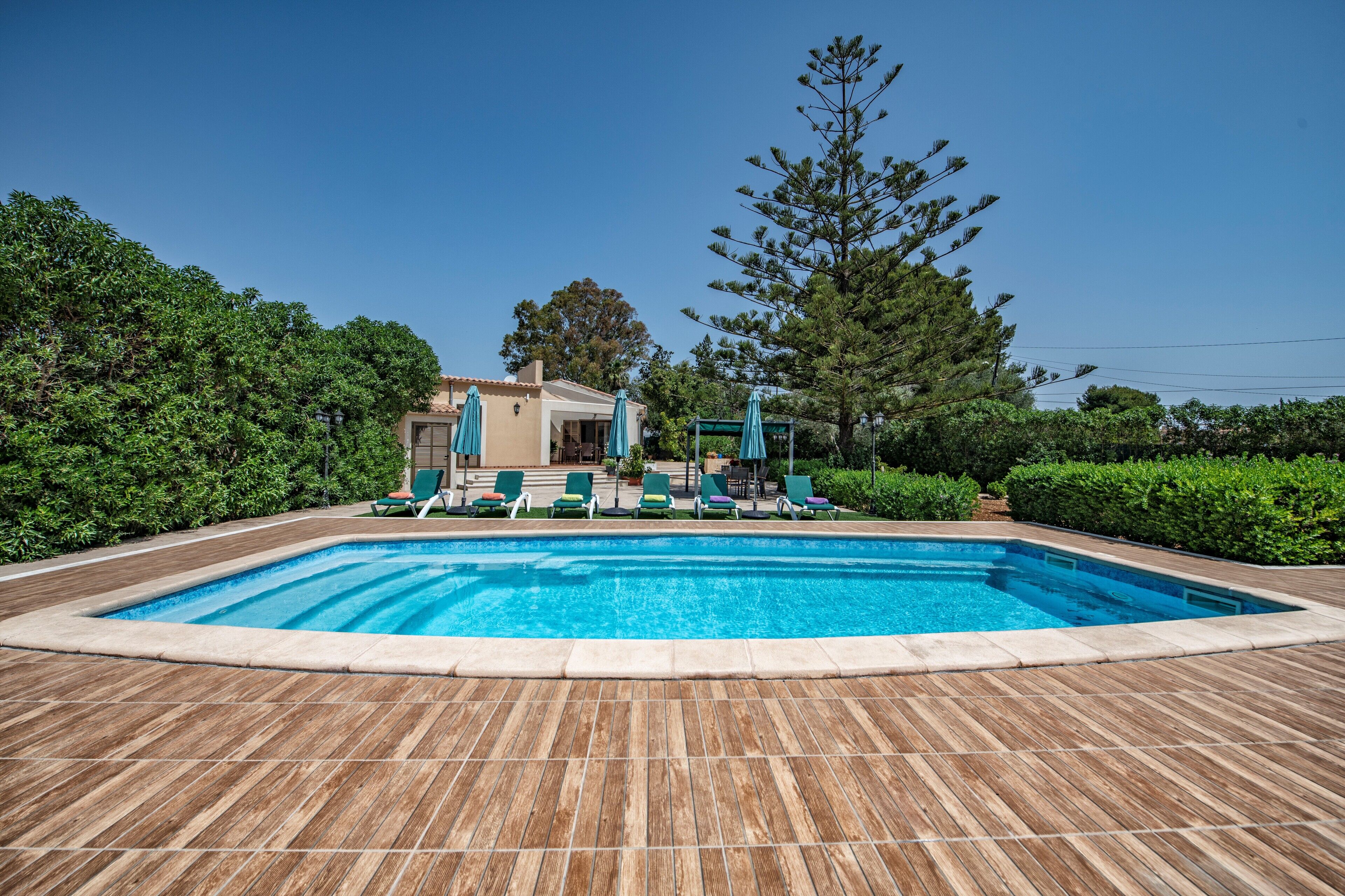 A rectangular swimming pool with a wooden deck, surrounded by green hedges and trees.