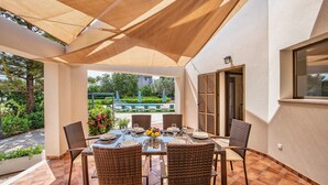 An outdoor patio with a table set for dining, surrounded by wicker chairs, and a shaded area with a view of a garden and buildings in the background. - HOUSE IN THE AREA OF CALA BONA WITH POOL AND GARDEN (CALA BONA)
