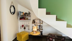 Un salon chaleureux avec un pouf jaune, une table ronde en bois surmontée d’une lampe, une bibliothèque remplie de livres et d’objets décoratifs, et une valise posée sur le sol.