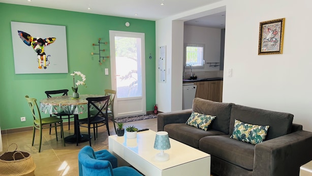 Un salon moderne avec un canapé gris, un fauteuil bleu et une table basse blanche. On y trouve également un coin repas avec une table et des chaises, et une cuisine en arrière-plan.