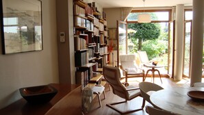 Ein helles Zimmer mit einer großen Bücherwand, einem Esstisch mit Stühlen und Blick ins Freie durch ein großes Fenster.