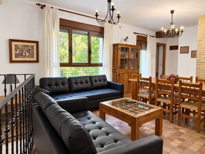 A living room with a sofa, a dining table, and chairs. There is a staircase with a railing, a chandelier, and a window with curtains. - Rural house Nº13 Molino Jaraiz for 8 people (Yeste)