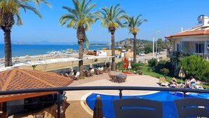 A poolside area with palm trees, a view of the sea, and a building with a tiled roof. - Poseidon 3, seafront 3 bedroom apartment with unobstructed beach and sea views. (Calis Plaji)