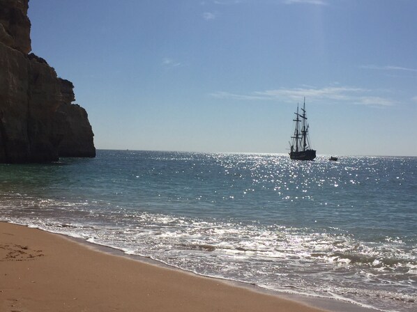 A beach with clear water, a ship, and a rocky cliff.