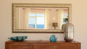 Un aparador de madera con un espejo grande encima, que refleja una ventana con cortinas y vistas al mar.