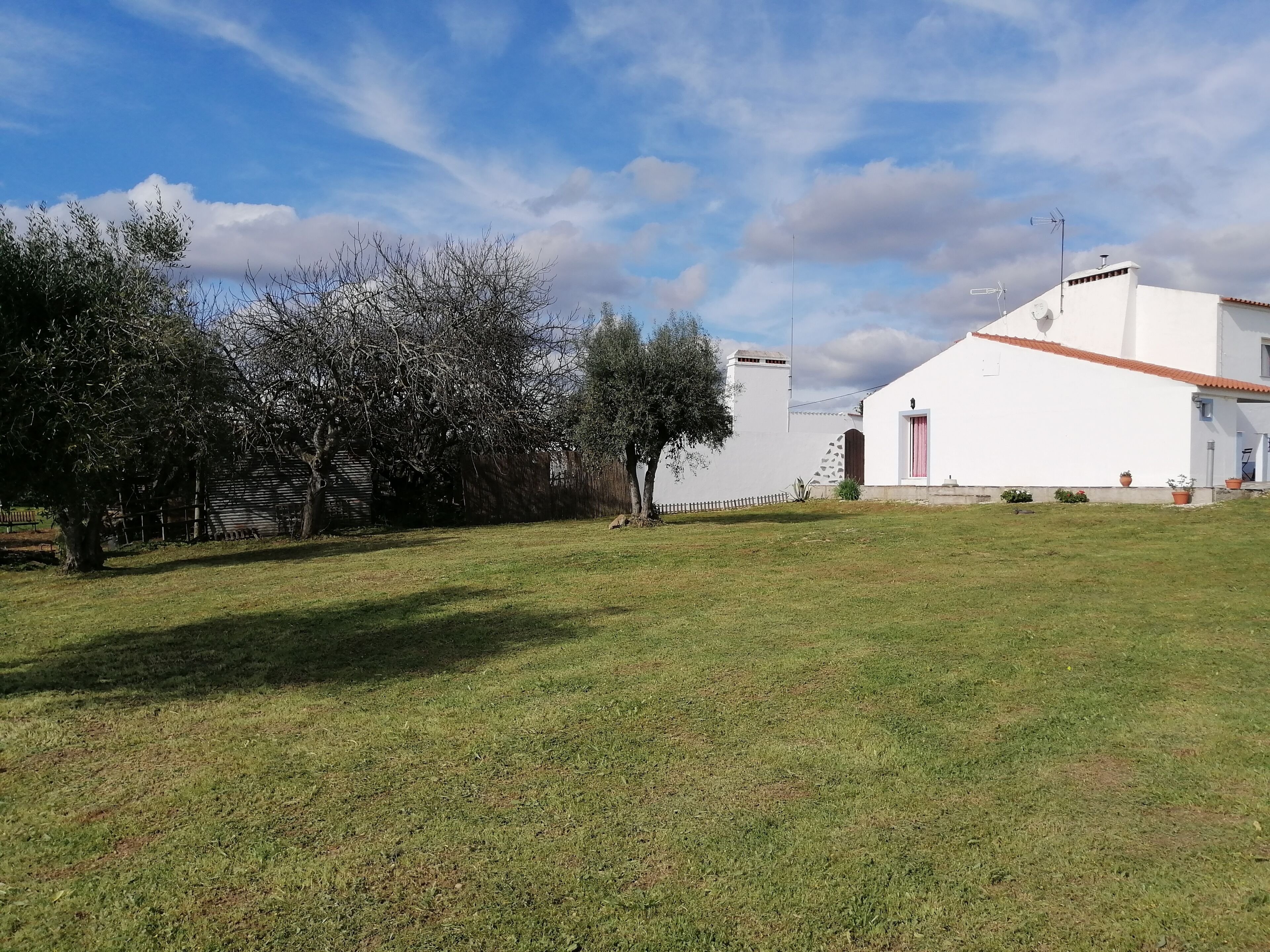 A white building with a red roof, a fenced area with trees, and a grassy field.